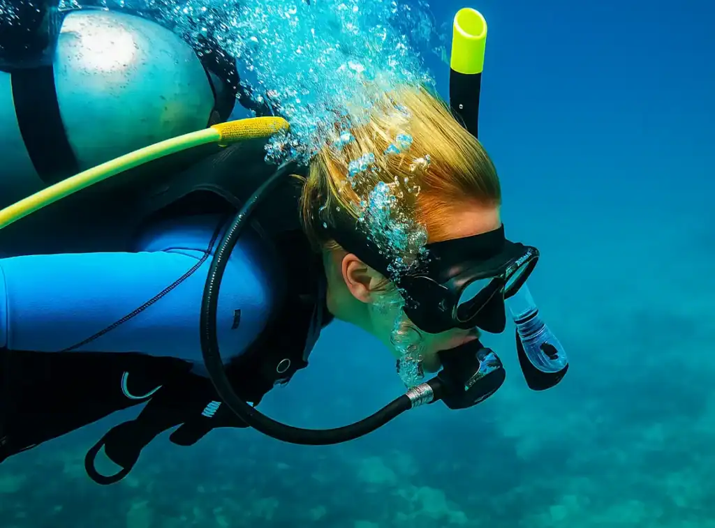 Dr Hayley Frend scube diving. She has on a wetsuit with a scuba diving mask on her face. On her back is a oxygen tank. A stream of bubbles rises from her respirator. Her blonde hair flows behind her.