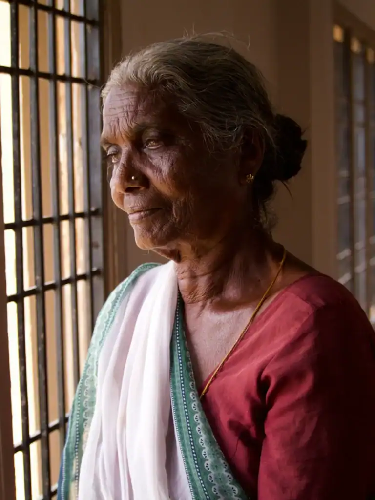 An elderly indian woman looks out of a window. The discolouration of her eyes suggests she is blind or partially sighted. She has a peaceful but resigned look on her face.  
