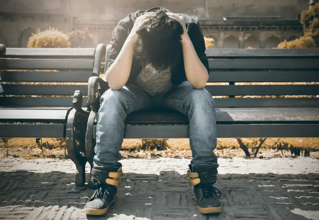 A young man sit on a bench his elbows on his knees and his hand wrapped around his bent head. He looks sad and in need of help. 