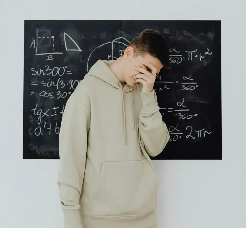 A teenage boy in a beige hooded jumper stands in front of a blackboard, which is filled with mathematical symbols and equations. The boy has his head in his hands in despair. It represents the frustration people with dyscalculia feel.