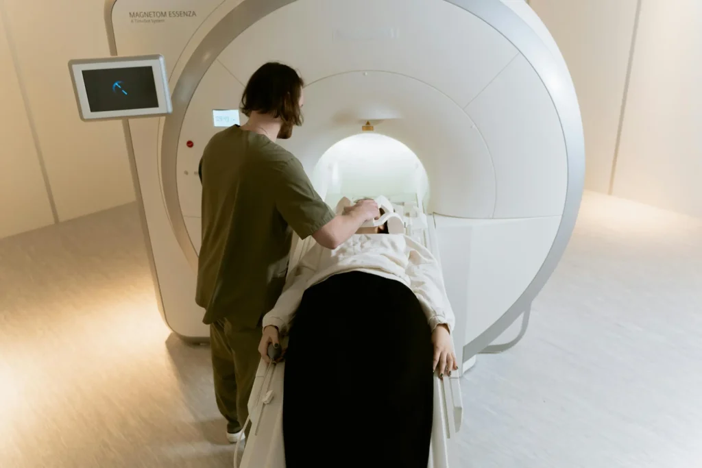 A patient lies on their back about to enter an MRI machine. A male nurse stands next to them. 