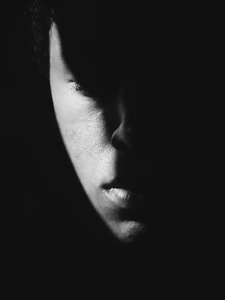 A face is half covered in dark shadow. The expression is somber giving a dark heavy feeling to the photo. It represents depression and bad mental health