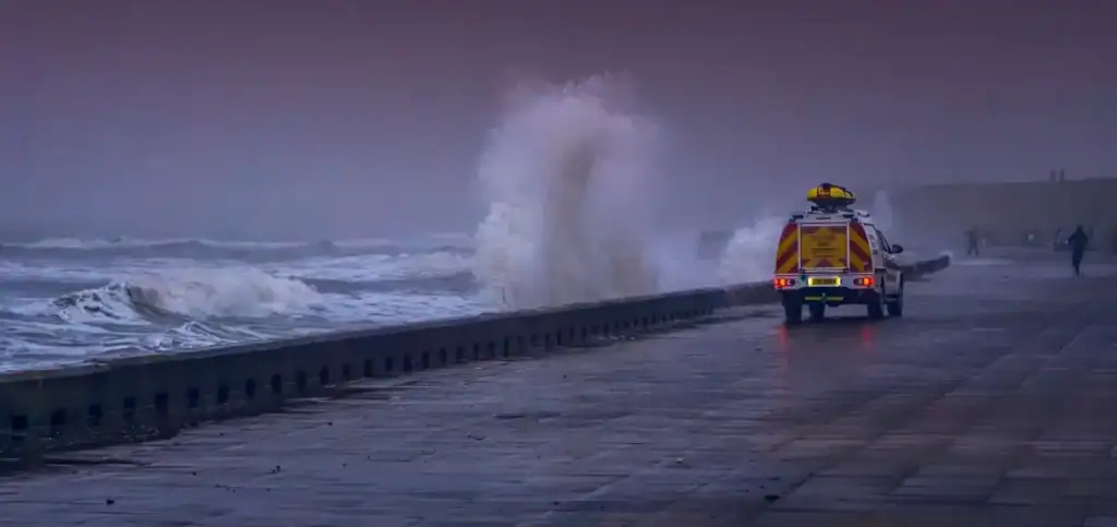 A low wall and beyond that a wild and stomry sea. A particularly large wave is breaking over the wall. The floor in front of the wall is wet, indicating the waves have been breaking over the wall for some time. a coastguard van with orange lights flashing is driving alongside the wall. The scene evokes the devastating power of storm waves and leaves an uneasy feeling.