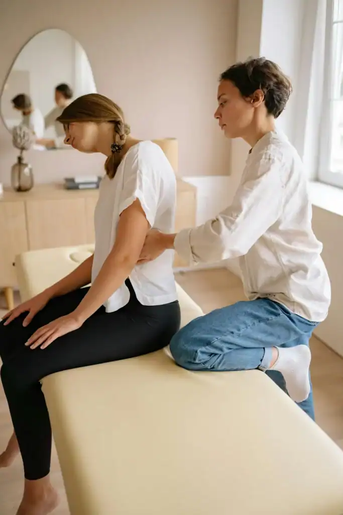 A woman sit on a massage table with her head cocked to one side while a physiotherapist kneels behind her and places her hand on the patients sides. 