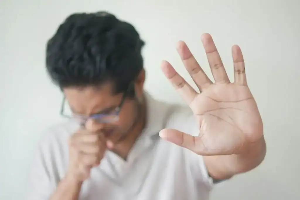 a man in a whote shirt holds his fist to his mouth as he is coughing. His other hand extends towards the camera, palm towards the camers in a 'stop' symbol. His forehead is creased, showing his apparent stuggle and suffering.