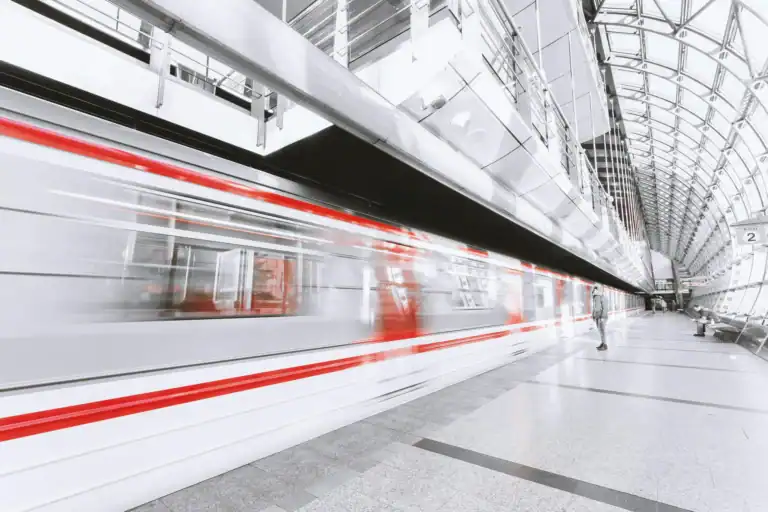 A train speeds through a modern looking station. Everything is white and clean, giving a futuristic feel. The train is blurred due to travelling so fast. It gives the impression of impressive speed.