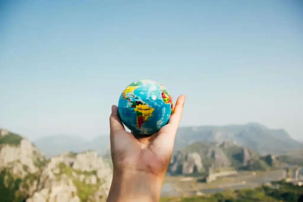 A hand is holding a small globe. In the background is a picturesque landscape of rugged mountains. It evokes the feeling of the world being small and ours to explore. 