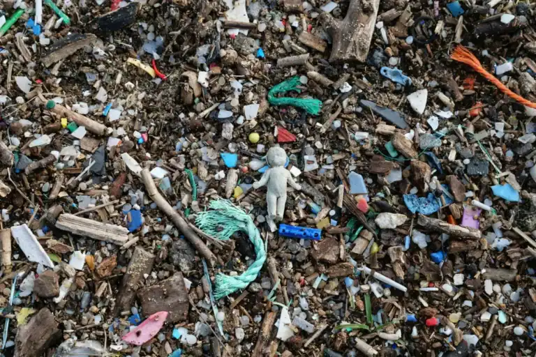 A stony beach floor is covered with countless pieces of plastic plastic. Many small shards of broken plastic and small pieces of string can be seen. At the center is a small white plastic figure of a cherub, its hand held up as if in despair.