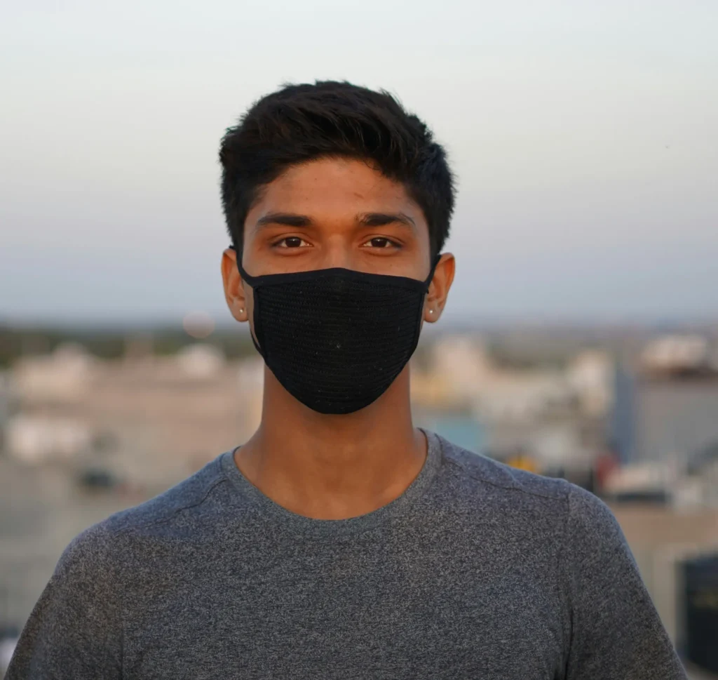 A man with short black hair stands looking at the camera. He is wearing a black face mask. A city can be seen behind him, stretching into the distance.