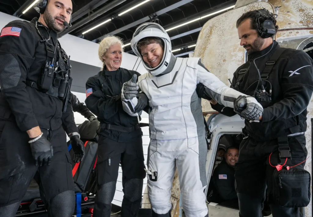 An astronaut wearing a white spacesuit smiles at the cameras and gives a thumbs-up. She is being supported by three people in black jumpsuits. It is clear she has just landed.