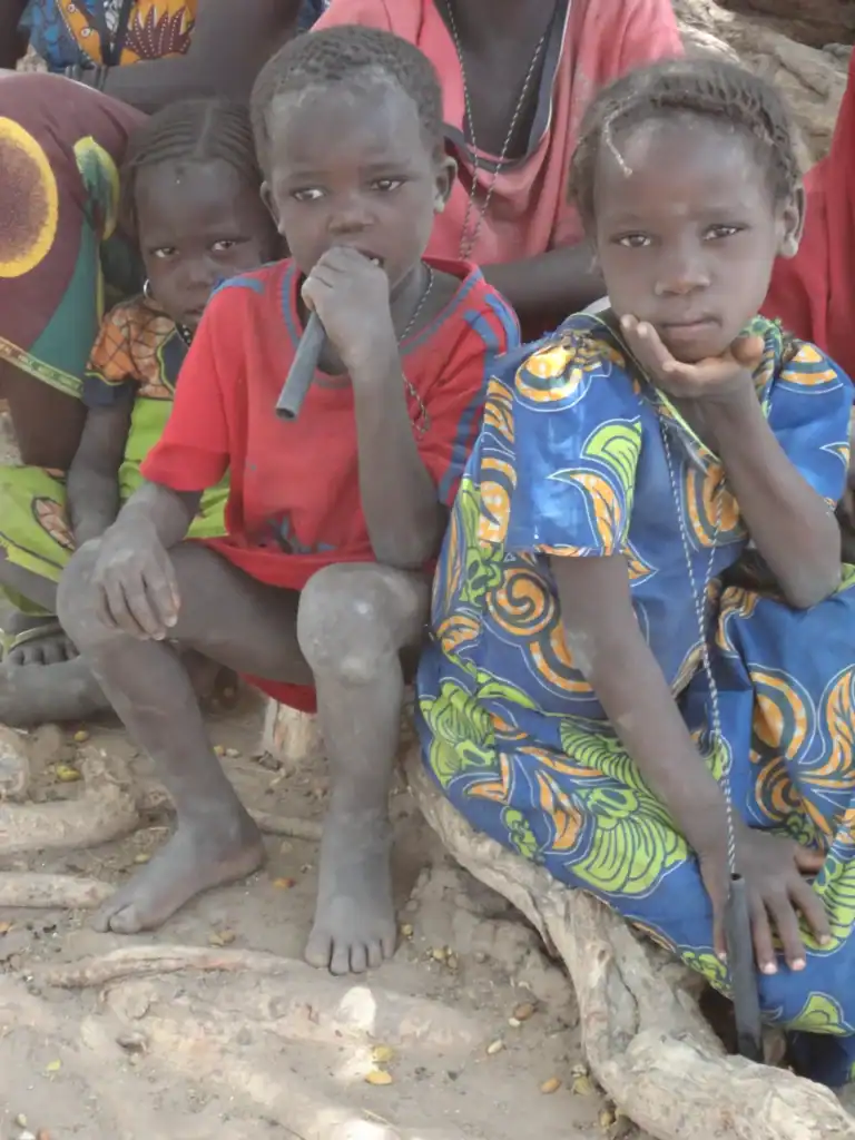 Children in Chad can be seen sat barefoot on the floor. A girl has a pipe hung from a chain around her neck. A boy has a long thin pipe in this mouth