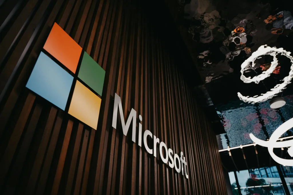 A illuninated sign on a wood panneled wall reads 'Microsoft' and the Microspft logo with its four coloured boxes arranged in a square is next to it. The wall appears to stand in a high tech office. 