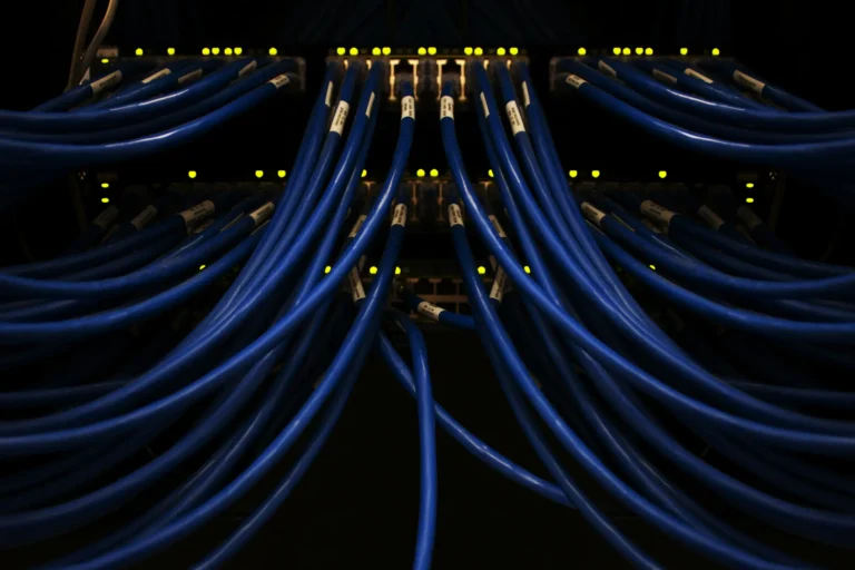 A large volumn of dark blue cables emergy from a darkened server with only numberous glowing lights visible. The cables extend out of the image in every direction.